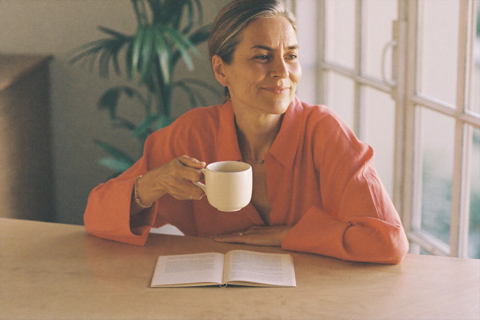 Formation abondance financière : femme sereine, tasse en main et carnet ouvert près d'une grande fenêtre lumineuse